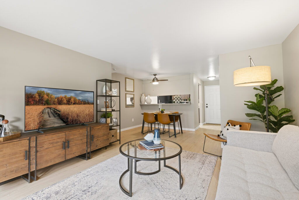 A modern living room with a light gray sofa, round glass coffee table, wooden TV stand, flat-screen TV, open shelving, dining area with four chairs, and a potted plant beside a floor lamp. Light walls and wooden flooring.