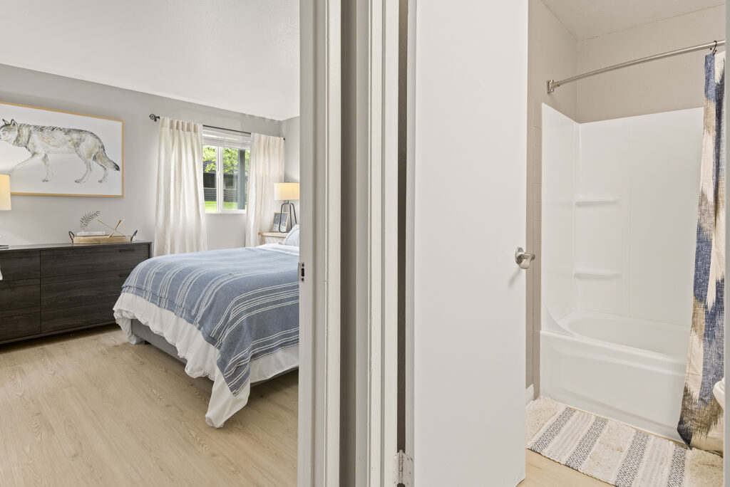 A view into a bedroom and bathroom; the bedroom has a bed with blue-striped bedding, a dresser, and a wolf picture on the wall, while the bathroom has a white bathtub with a patterned shower curtain.