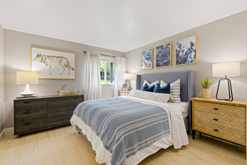 A cozy bedroom with a blue-striped bed, two bedside tables with lamps, a dresser, wall art including a wolf print, and a window with sheer curtains letting in natural light.