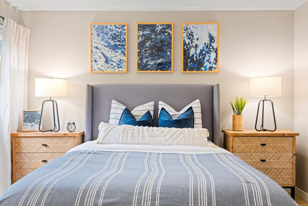 A modern bedroom with a gray upholstered bed, blue and white bedding, two wooden nightstands with lamps, a potted plant, and three abstract blue artworks above the bed. Light enters from a window on the left.