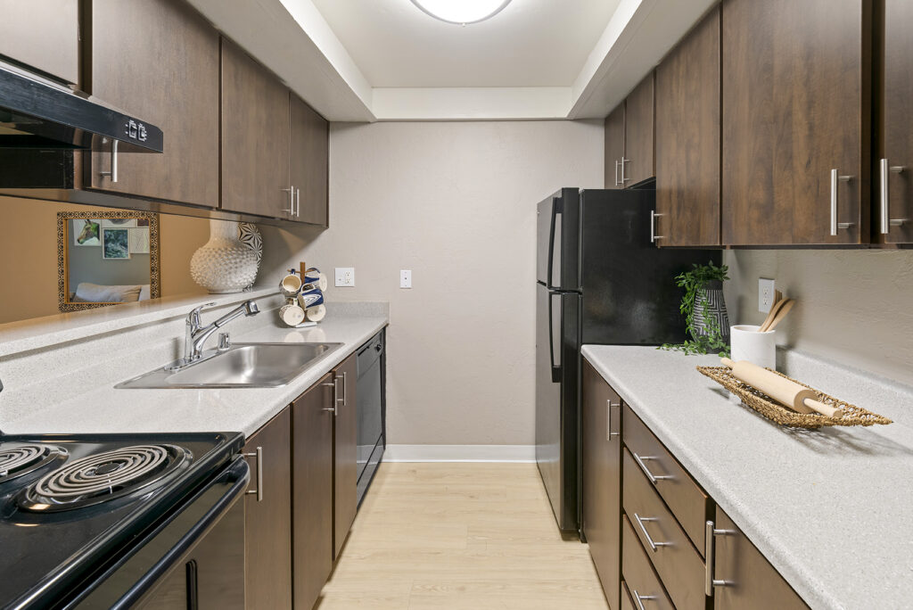 Modern galley kitchen with dark wood cabinets, light countertops, black appliances, a double sink, electric stove, and decorative items on the counters. A mirror and vase are visible in the adjacent room.