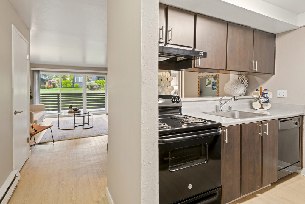 A modern apartment kitchen with dark wood cabinets and black appliances is adjacent to a bright living room with large windows, a beige sofa, and a coffee table on a light wood floor.