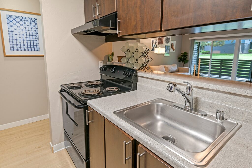 Small kitchen with a stainless steel sink, electric stove, dark wood cabinets, and a light countertop. Beyond the kitchen, there is a living area with a window showing greenery outside.