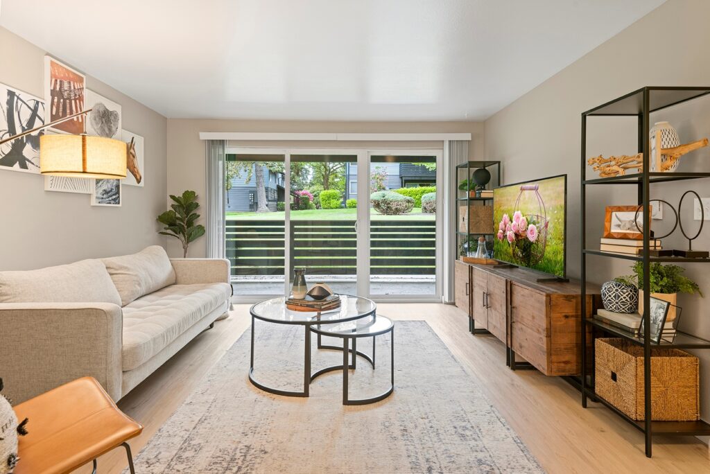 Modern living room with a beige sofa, round glass coffee tables, TV on a wooden stand, open shelves, and large sliding glass doors revealing a green, landscaped outdoor area with grass and shrubs.