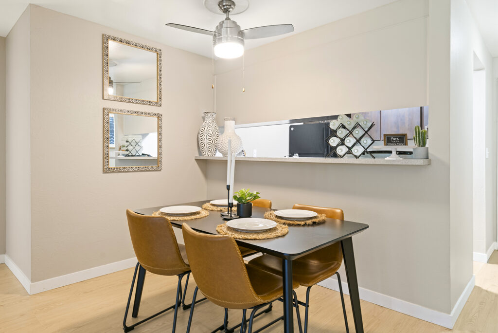 A modern dining area with a black table set for four, tan leather chairs, woven placemats, and a small plant centerpiece. Two decorative mirrors hang on a beige wall, and a ceiling fan light is overhead.