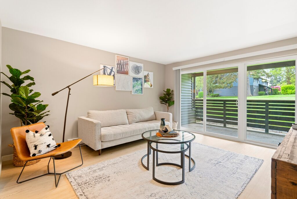 A modern living room with a beige sofa, a tan leather chair, a round glass coffee table, potted plants, wall art, and large sliding glass doors opening to a patio and green outdoor space.