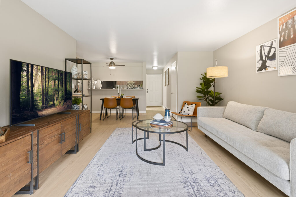 Modern living room with a light gray sofa, glass coffee table, wooden TV stand, large flat screen TV, dining area with four orange chairs, and kitchen in the background. Neutral tones and minimalistic decor throughout.